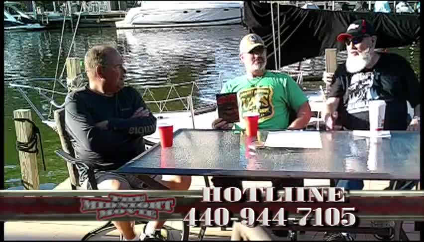 Three men sitting around a glass table on a dock. Water and boats behind them. 