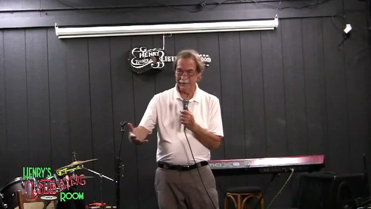 Black paneled wall in the back. Man in button up shirt holding a mic in the middle.