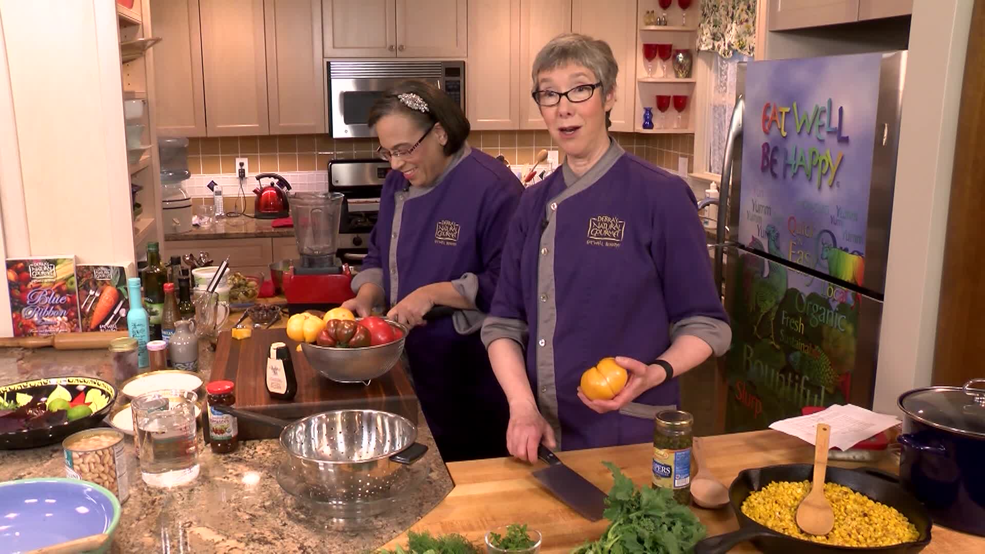 Kitchen setting. Woman with glasses and purple chef jacket to the middle right. Woman with dark hair and purple chef jacket to middle left preparing food.