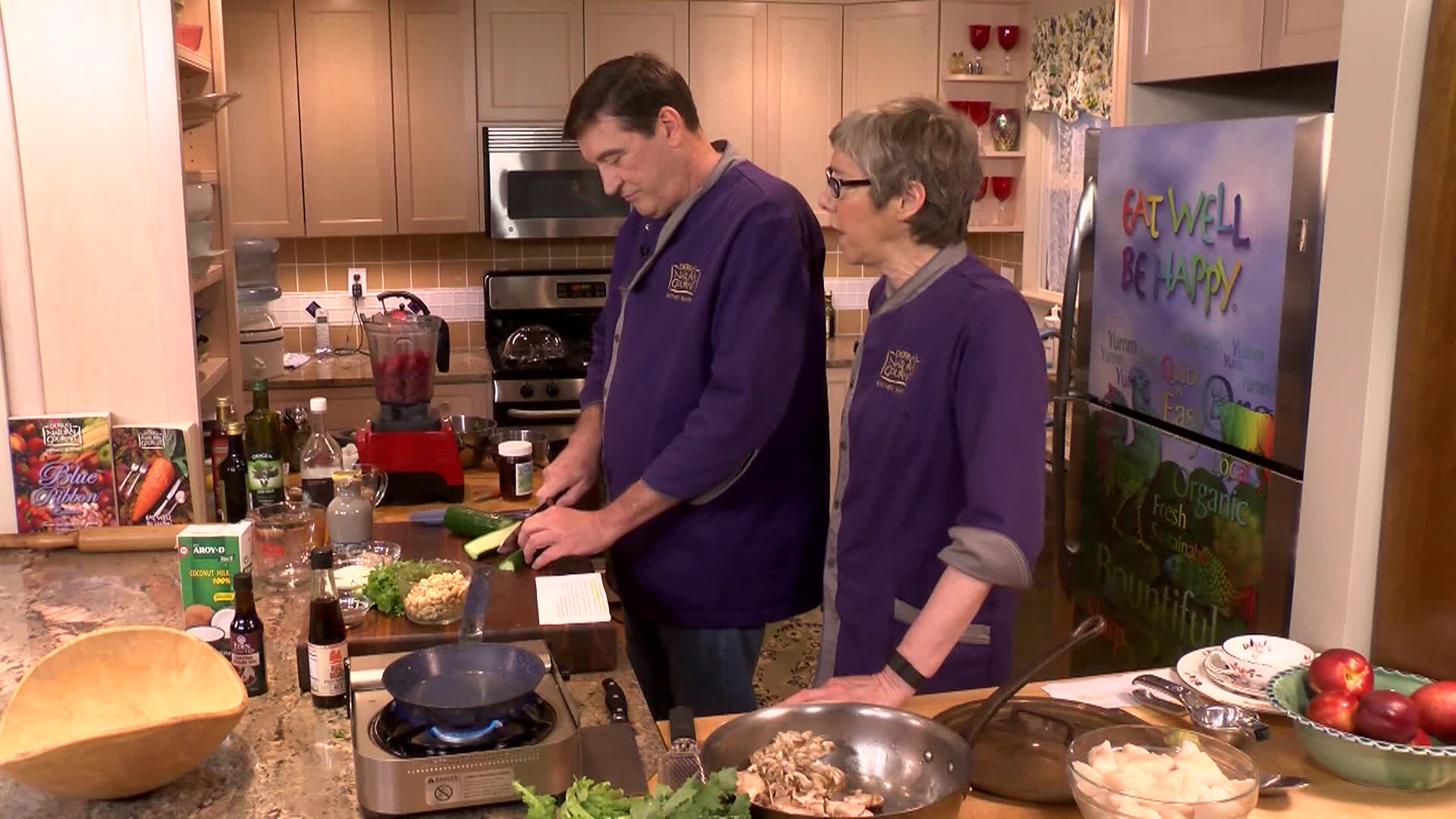 Kitchen setting. Woman with glasses and purple chef jacket to the middle right. Man with glasses and purple chef jacket to middle left preparing food.