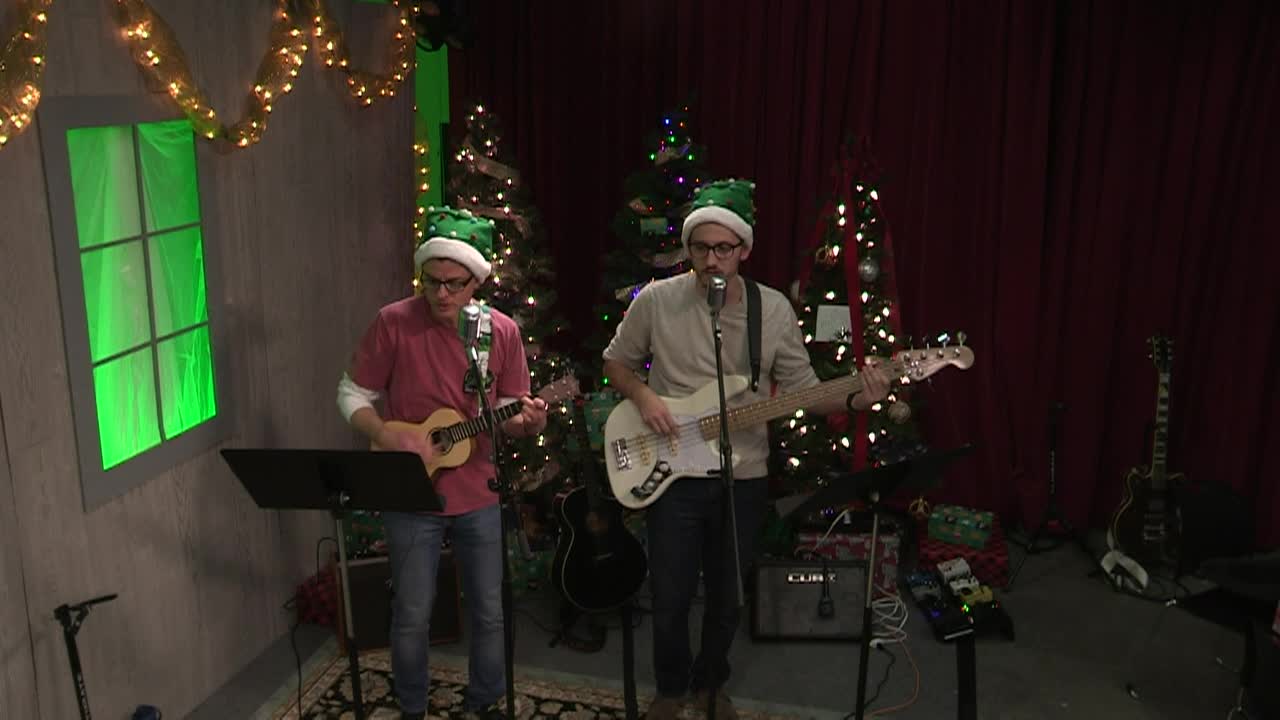 Holiday scene. Man in red and Santa hat playing a ukulele in front of a mic to the left. Man next to the other in tan, wearing a santa hat playing guitar  