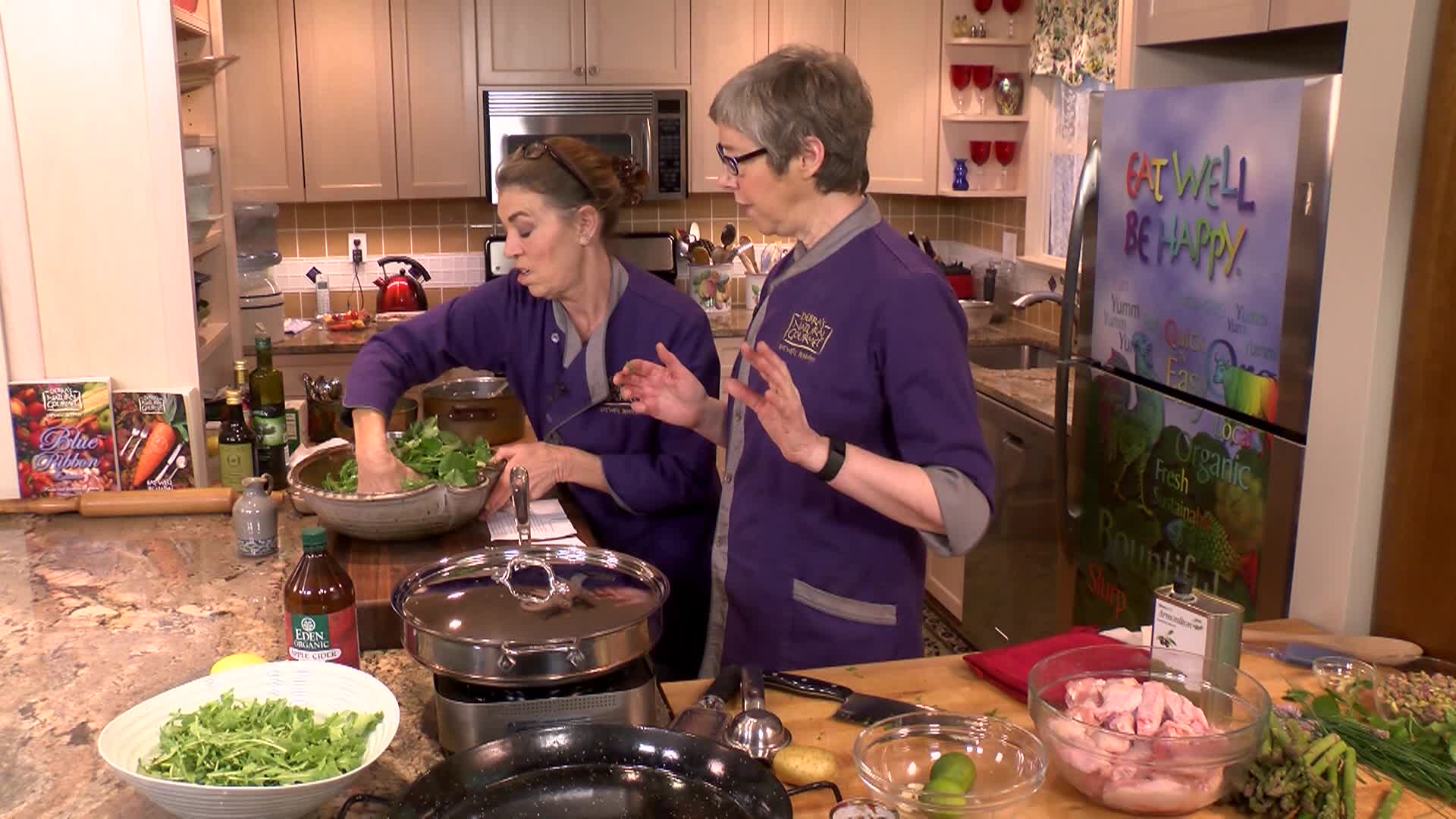 Woman in purple on the left in a kitchen cooking. Woman in purple in the center prepping food.